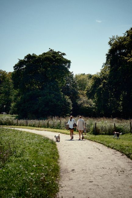 Two women walking along a curved gravel pathway in a well-maintained, lush communal garden on the Isle of Dogs, with one carrying a bag and both dressed in light clothing suitable for mild weather. Th